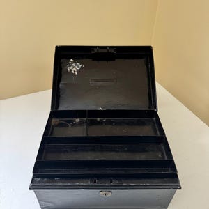 Puede incluir: Caja registradora de metal negro abierta con un ojo de cerradura y compartimentos interiores. La caja rectangular tiene una tapa con bisagras y una cerradura. El interior tiene varios compartimentos para organizar objetos.