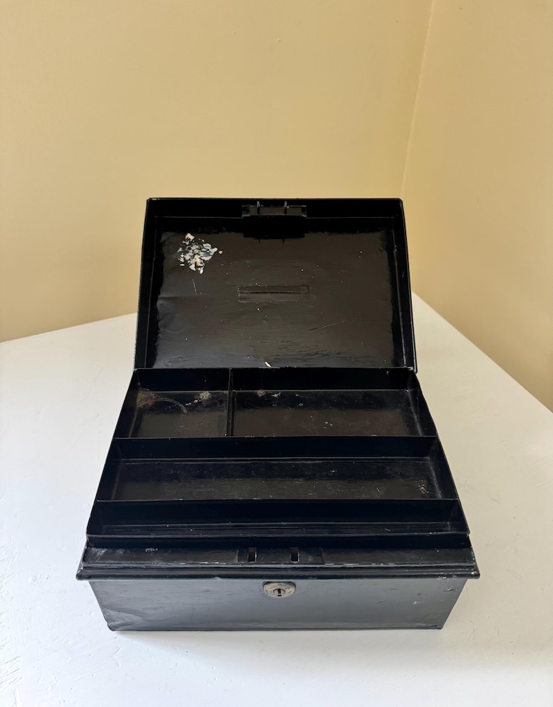 Puede incluir: Caja registradora de metal negro abierta con un ojo de cerradura y compartimentos interiores. La caja rectangular tiene una tapa con bisagras y una cerradura. El interior tiene varios compartimentos para organizar objetos.