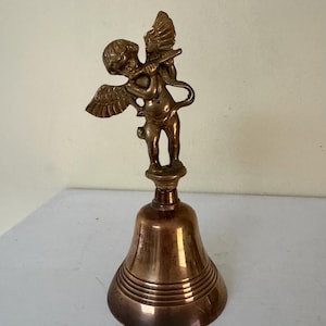 May include: A bronze-colored bell with a cherub figurine playing a flute on top. The cherub has wings and is standing on a decorative base. The bell has horizontal ridges around its base.