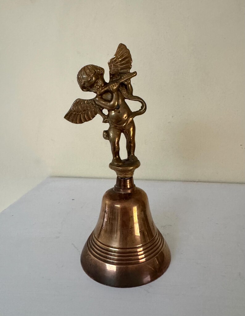 May include: A bronze-colored bell with a cherub figurine playing a flute on top. The cherub has wings and is standing on a decorative base. The bell has horizontal ridges around its base.