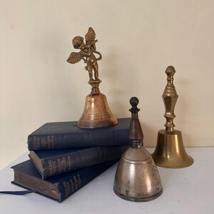 May include: An assortment of antique bells, including a bronze bell with an angel figurine, a silver-toned bell with a wooden handle, and a brass bell. The bells are displayed on a stack of blue books.