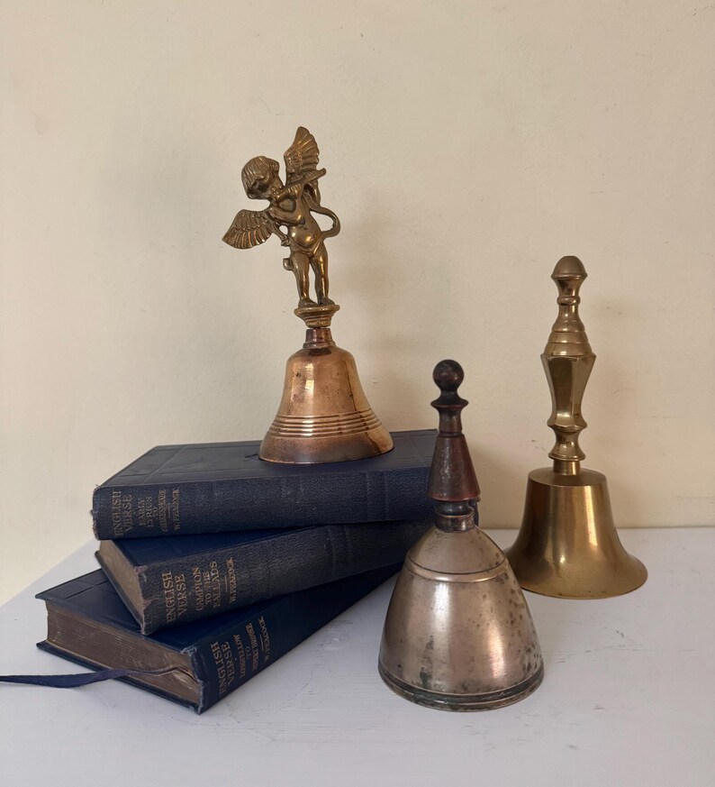 May include: An assortment of antique bells, including a bronze bell with an angel figurine, a silver-toned bell with a wooden handle, and a brass bell. The bells are displayed on a stack of blue books.
