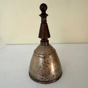 May include: A vintage hand bell with a tarnished metal body and a dark wooden handle. The bell has a conical shape with a rounded top and a dark wooden handle. The handle is composed of a small ball, a conical section, and a cylindrical base.