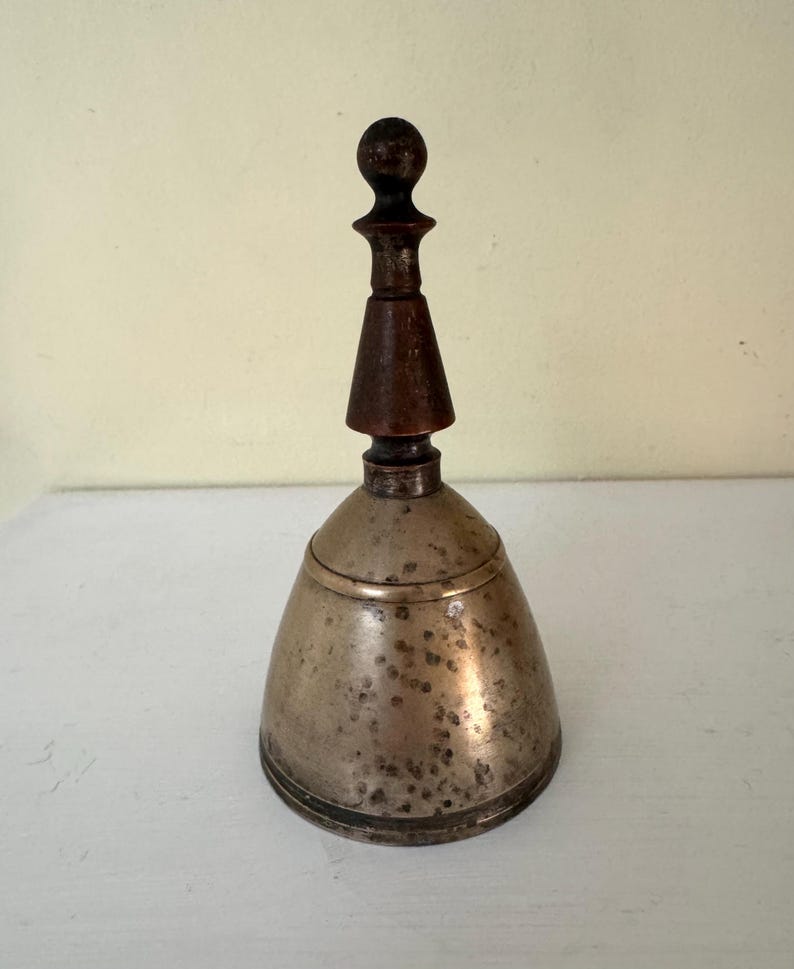May include: A vintage hand bell with a tarnished metal body and a dark wooden handle. The bell has a conical shape with a rounded top and a dark wooden handle. The handle is composed of a small ball, a conical section, and a cylindrical base.