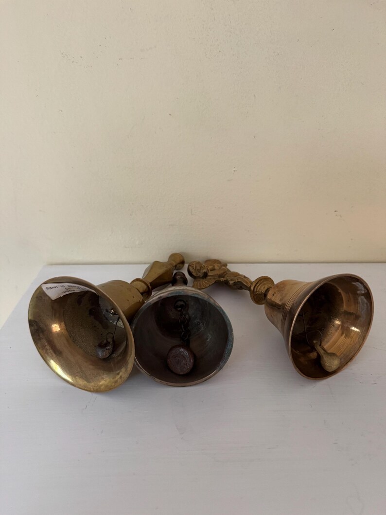 May include: Four antique brass bells with varying handle designs. The bells are arranged on a white surface, showcasing their aged, golden appearance. The bells have a classic, rounded shape, and the handles feature decorative elements.
