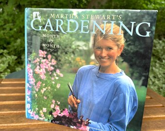 May include: A hardcover book titled Martha Stewart's Gardening Month by Month. The cover features a photograph of a person in a blue long-sleeved shirt holding garden shears in a garden setting with pink flowers. The text is printed in white.