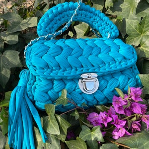Puede incluir: Un bolso de mano de crochet azul turquesa con una correa de cadena plateada y un borla a juego. El bolso tiene un cierre de solapa con un broche plateado.