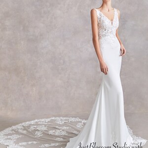 May include: A white wedding dress with a fitted bodice and a flowing skirt. The dress features delicate lace detailing and a V-neckline. The dress is paired with a long, flowing veil. JustBlossomStudio with Adela Designs.