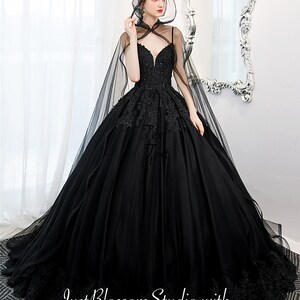 May include: A black tulle ball gown with a lace bodice and a long black tulle cape. The gown has a sweetheart neckline and a fitted bodice. The skirt is full and flowing. The cape has a hood and is trimmed with lace.