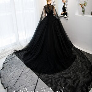May include: A black tulle ball gown with a high neck and a cape. The gown has a fitted bodice and a full skirt. The model is standing in front of a mirror, and the gown is flowing behind her.