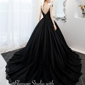 May include: A black tulle ball gown with a lace bodice and a deep V-neckline. The gown has a long train and is decorated with floral lace appliques. The gown is shown on a model in a white room with a mirror.