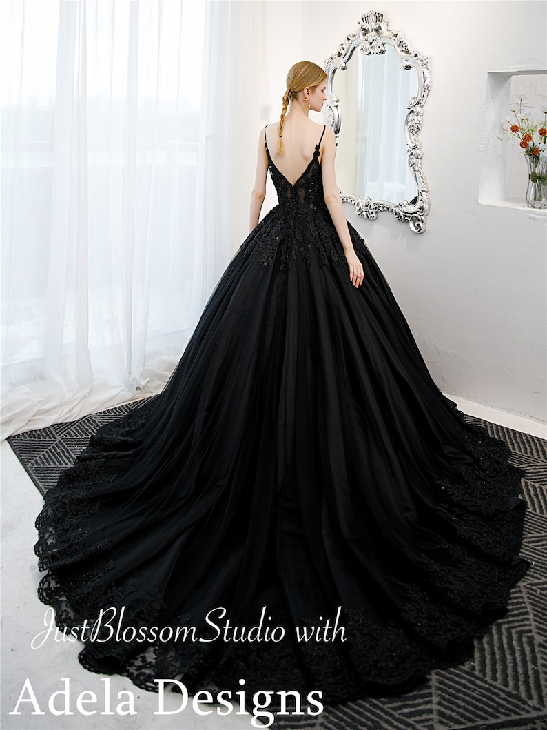 May include: A black tulle ball gown with a lace bodice and a deep V-neckline. The gown has a long train and is decorated with floral lace appliques. The gown is shown on a model in a white room with a mirror.