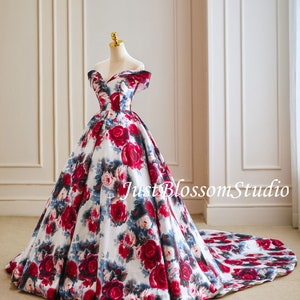 May include: A white off-the-shoulder ball gown with a red and blue floral print. The gown has a full skirt and a long train. The dress is on a mannequin in a room with white walls.