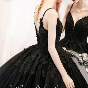 May include: A black tulle ball gown with a lace overlay and a plunging neckline. The dress features a fitted bodice and a full skirt. The dress is shown on a model with a braided hairstyle. The text "Just Blossom Studio with Adela Designs" is visible at the bottom of the image.