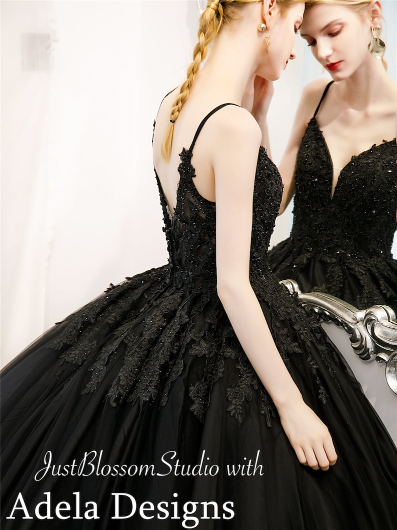 May include: A black tulle ball gown with a lace overlay and a plunging neckline. The dress features a fitted bodice and a full skirt. The dress is shown on a model with a braided hairstyle. The text "Just Blossom Studio with Adela Designs" is visible at the bottom of the image.