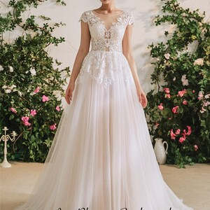 May include: A white tulle wedding dress with lace detailing and a sheer bodice. The dress has a scoop neckline and short sleeves. The model is standing in front of a floral backdrop.