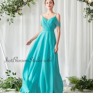 May include: A turquoise chiffon gown with a V-neck and off-the-shoulder sleeves. The dress has a flowing skirt, suitable for formal events. The text "JustBlossom Studio with Adela Designs" is visible.