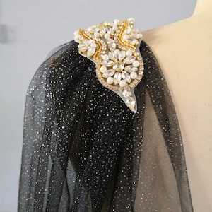 May include: Black sheer fabric with silver glitter and a gold and white beaded embellishment on the shoulder.