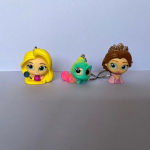 May include: Three character keychains: a blonde princess with a mirror, a green chameleon, and a brunette princess with a crown. Each figure has large eyes and is attached to a silver keyring. The figures are made of plastic.