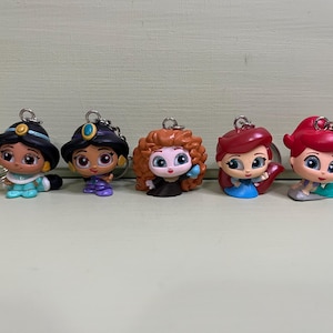 May include: Five colorful character keychains featuring Jasmine, a princess in a black hat, another princess in a purple dress, Merida with red curly hair, Ariel with red hair, and Ariel with red hair. Each keychain has a silver ring.