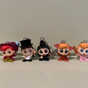 Puede incluir: Cinco llaveros de personajes con anillos de metal plateado. Las figuras son de estilo caricatura, con ojos grandes y varios atuendos. Los personajes incluyen una figura con un atuendo rojo y morado, una figura con un sombrero de copa negro y una figura con una capa negra.