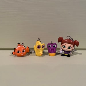 May include: Four character keyrings from the animated film, "Finding Nemo." The keyrings feature Nemo, Dory, a purple character, and a girl with red hair. Each keyring has a silver ring for attaching to keys or bags.