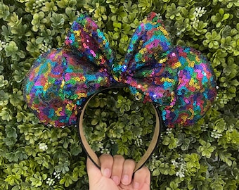 Op de afbeelding: Een paar kleurrijke, met pailletten bezette Minnie Mouse oren met een grote strik. De strik is gemaakt van iriserende pailletten in tinten roze, blauw, groen en paars. De oren zijn zwart en hebben een bruin hoofdband.