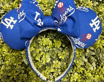 May include: A blue headband featuring two round ears and a central bow, all covered in a repeating pattern of the white Dodgers logo, the word Dodgers in white script, and small red and white baseball graphics with motion lines.