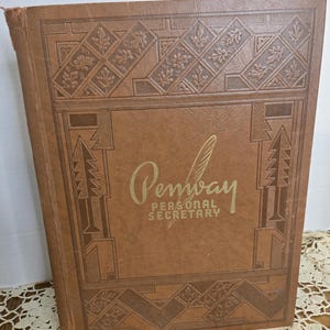 Pode incluir: Um livro vintage castanho "Pembay Personal Secretary". A capa apresenta desenhos geométricos e florais em relevo. O título está em letras douradas. O livro está ligeiramente desgastado, sugerindo idade e uso. A estética geral é clássica e funcional.