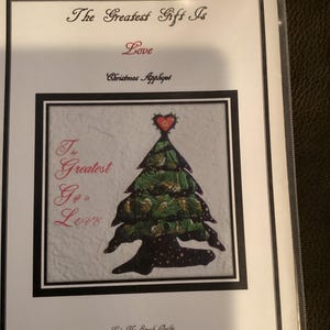Peut inclure: Un motif d'appliqué de Noël représentant un arbre vert et or avec un cœur rouge au sommet. Le texte "The Greatest Gift Is Love" est écrit en rose sur fond blanc. Le motif est intitulé "Christmas Appliqué" et est réalisé par "Let Me Speak Quilts".