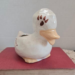 Może przedstawiać: Ceramiczna doniczka w kształcie kaczki w kremowobiałym kolorze z jasnobrązowym dziobem i stopami. Kaczka ma brązowe ślady łap na głowie i małe brązowe kropki z boku. Doniczka stoi na czerwonej powierzchni.