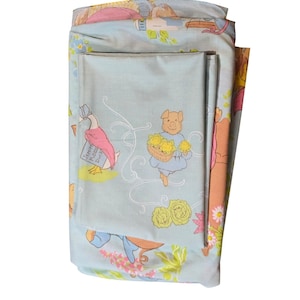 May include: Folded light blue bedsheets with a whimsical pattern featuring cartoon characters, including a pig carrying flowers and a duck holding a book. The sheets have a floral and scroll design with pink, yellow, and green accents.
