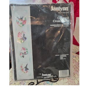 May include: A Janlynn counted cross-stitch kit featuring a design of hummingbirds and flowers. The kit includes 14-count Aida fabric and cotton floss. Finished size is 6 x 2.5 inches.
