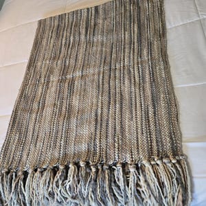 Puede incluir: Una manta rectangular tejida con un borde con flecos. La manta presenta rayas verticales en tonos beige, marrón y gris. La franja es una mezcla de hilos beige y grises, que añade textura e interés visual al diseño.