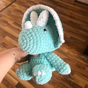 Könnte beinhalten: Ein gehäkeltes türkisfarbenes Triceratops-Dinosaurier-Plüschtier mit weißen Akzenten. Das Spielzeug hat einen weißen gehäkelten Kragen um den Kopf und weiße gehäkelte Hörner.
