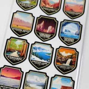 May include: A set of 12 colorful stickers featuring national parks in the United States. Each sticker is shaped like a shield and features a different park's name and a scenic image of the park. The stickers are perfect for adding a touch of nature to your belongings.