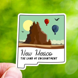 May include: A sticker featuring a stylized illustration of a mountain range with a sun and hot air balloons in the sky. The text "New Mexico The Land of Enchantment" is printed below the image.
