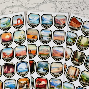 May include: A set of 48 stickers featuring colorful illustrations of national parks in the United States. Each sticker is shaped like a shield and features the name of the park and a scenic image. The stickers are printed on white paper.