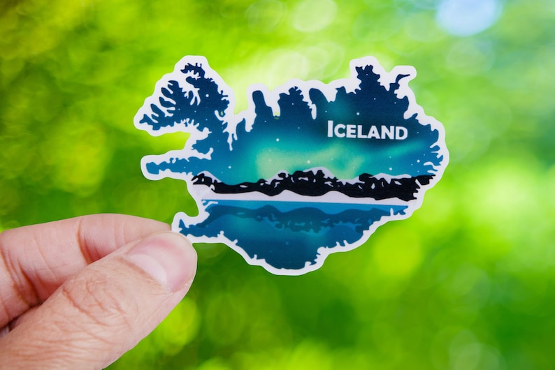 May include: An Iceland-shaped sticker with a scenic design. The design features the Northern Lights and a body of water, with the word "Iceland" in white. The background is green.