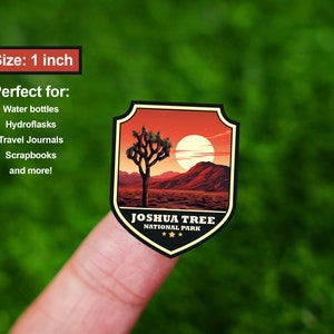 May include: A 1 inch sticker featuring a Joshua Tree National Park design. The sticker has a shield shape with a red, orange, and yellow sunset background. A silhouette of a Joshua Tree is in the center of the sticker. The text "Joshua Tree National Park" is printed in black at the bottom of the sticker.