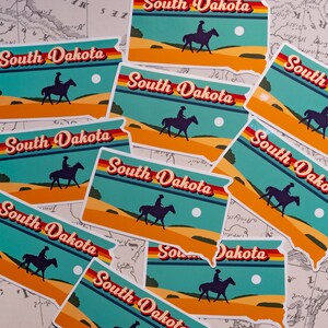 May include: A collection of nine colorful stickers featuring the state outline of South Dakota with the text "South Dakota" in a retro font. Each sticker features a silhouette of a person riding a horse across a desert landscape with a sun and clouds in the background.