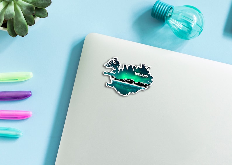 May include: An Iceland-shaped sticker with a scenic illustration of the Northern Lights. The design features green, blue, and black hues, with the word "Iceland" in white. The sticker is placed on a white laptop.