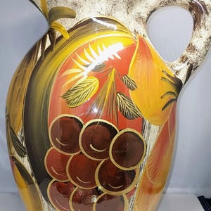 Puede incluir: Una jarra de cerámica con un diseño pintado a mano de uvas de color marrón oscuro y frutas amarillas sobre un fondo naranja cálido y crema. El recipiente tiene un asa ancha, un borde acampanado de color marrón oscuro y un acabado texturizado.