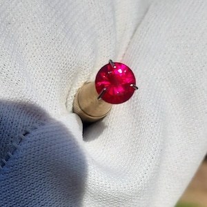 May include: A round, faceted, vibrant red gemstone set in a simple metal prong setting. The stone displays a brilliant, reflective surface with deep crimson hues, securely held by four thin metal prongs atop a tapered, metallic base component.
