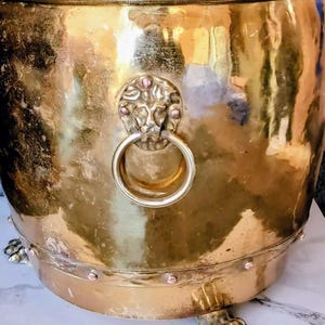 Puede incluir: Un cubo de latón pulido con un asa en forma de cabeza de león con un anillo circular, una banda base reforzada con remaches visibles y tres pies en forma de garra. La superficie metálica muestra un tono dorado cálido y reflectante.