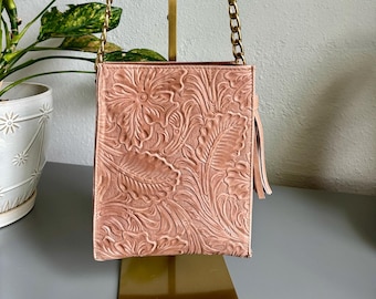 May include: A blush-colored handbag with a floral embossed design. The bag features a brown leather handle and gold chain straps. A small tassel hangs from the side. The bag is displayed on a gold stand.