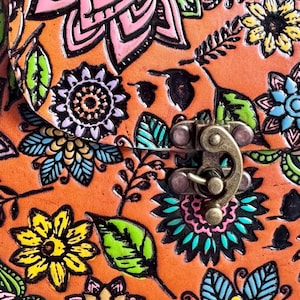 May include: Close-up of a leather bag with a floral design. The bag is orange with embossed flowers in pink, yellow, blue, and green. The bag has a metal clasp.