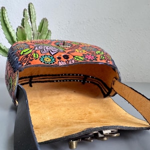 May include: Open leather crossbody bag with a vibrant floral pattern in orange, green, pink, and blue. The bag features a black strap and a brass clasp. The interior is a light tan colour, and the bag is open, revealing the inside.