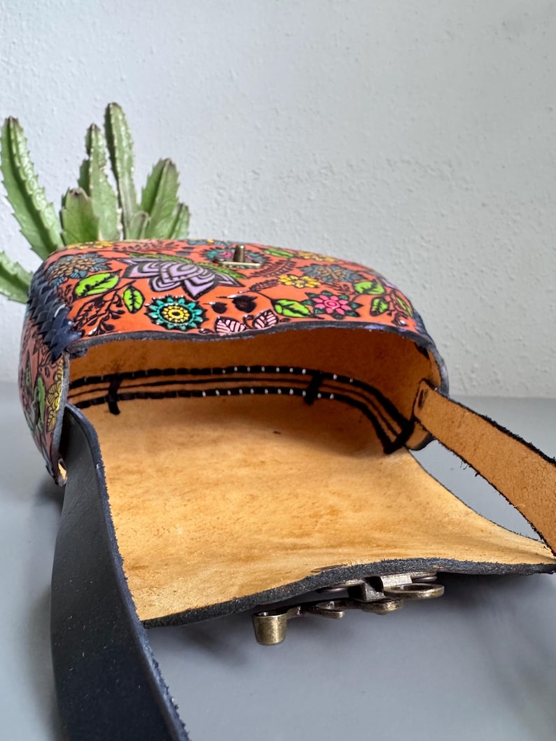 May include: Open leather crossbody bag with a vibrant floral pattern in orange, green, pink, and blue. The bag features a black strap and a brass clasp. The interior is a light tan colour, and the bag is open, revealing the inside.
