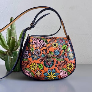 May include: A leather crossbody bag in orange, decorated with a floral pattern in pink, blue, yellow, and green. It features a dark blue strap and trim, and a metal clasp. A cactus plant is in the background.